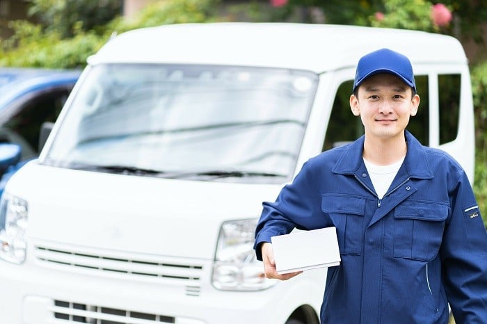 白い貨物軽自動車の前に、青い作業服を着て荷物を持った男性がいる