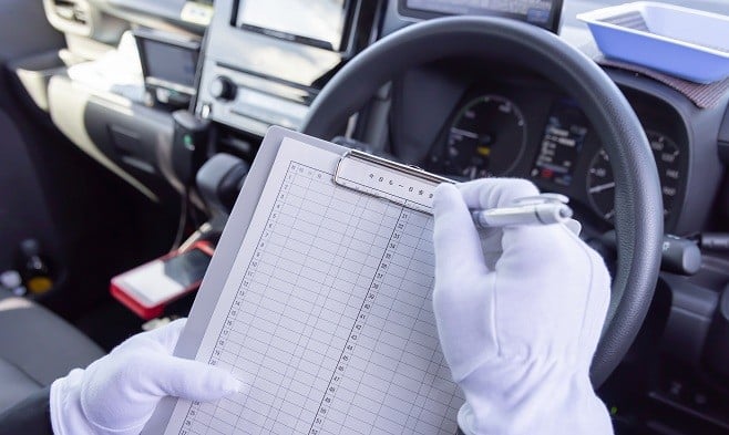 白い手袋をはめたドライバーが、車内でクリップボードの運転日報に記入している様子